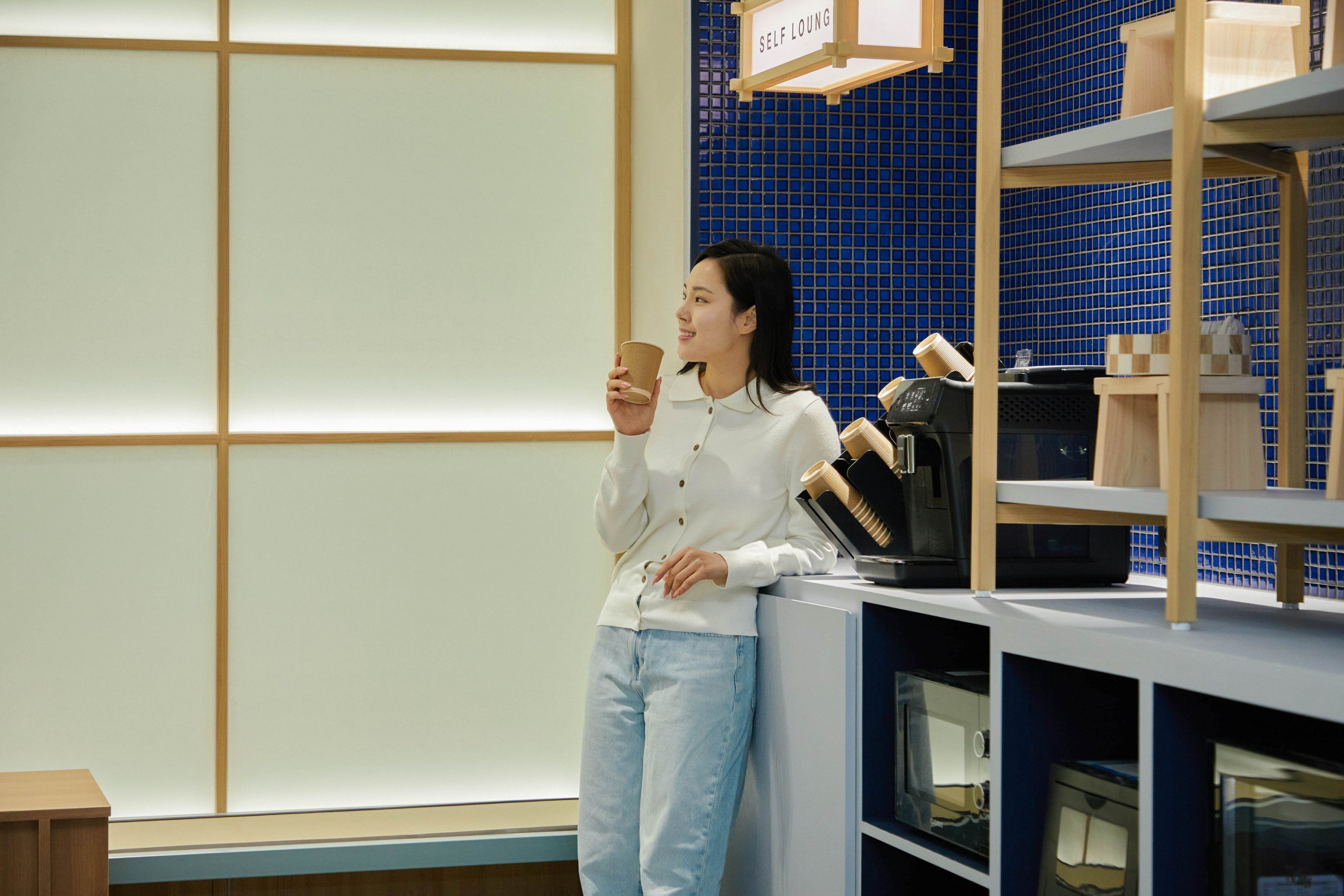 A person standing in a room with a blue tiled wall, holding a cup. There is a sign above that reads "SELF LOUNGE".