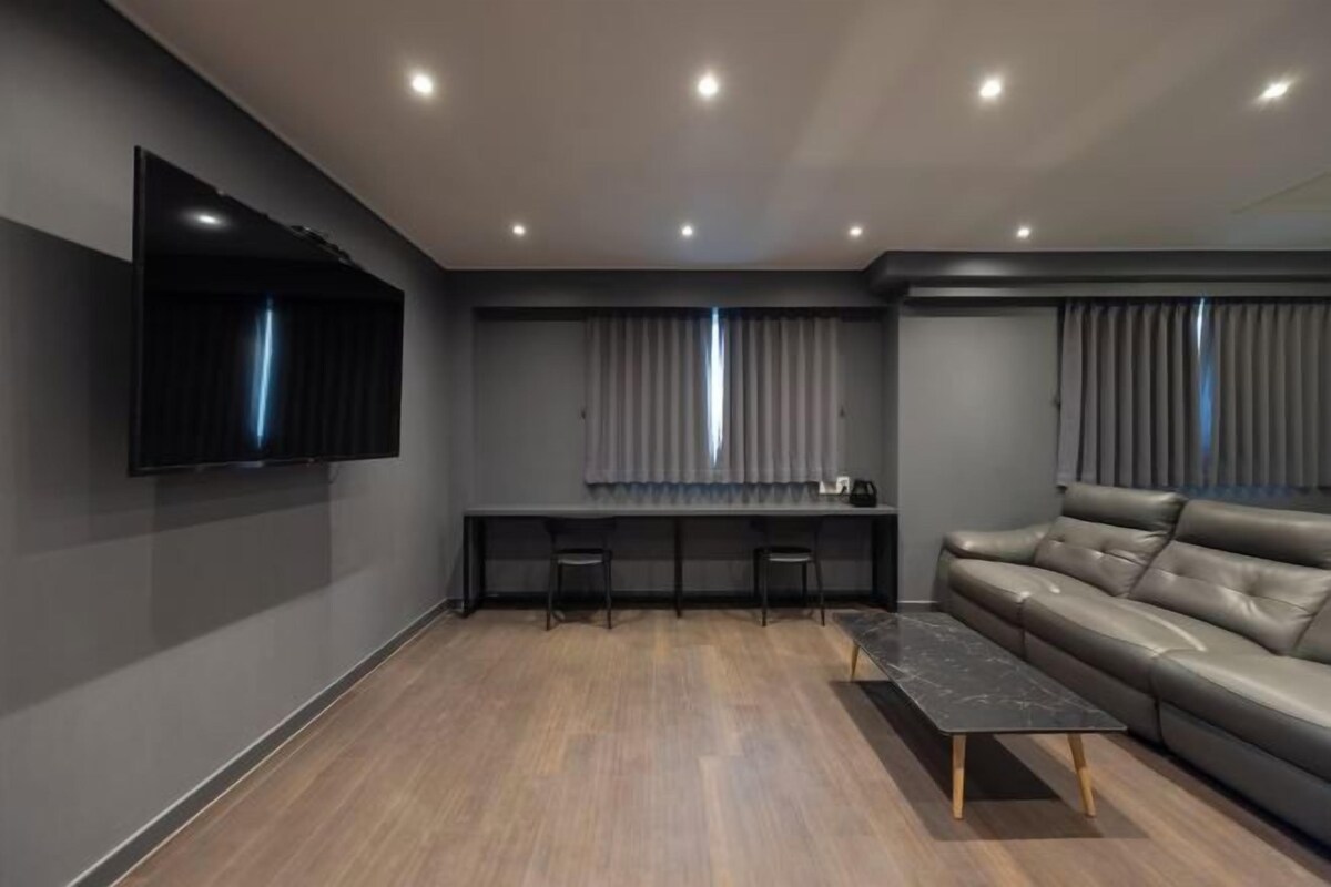 Una sala moderna con una televisión de pantalla plana montada en la pared, un sofá oscuro, una mesa de centro y un comedor con una mesa larga y sillas.