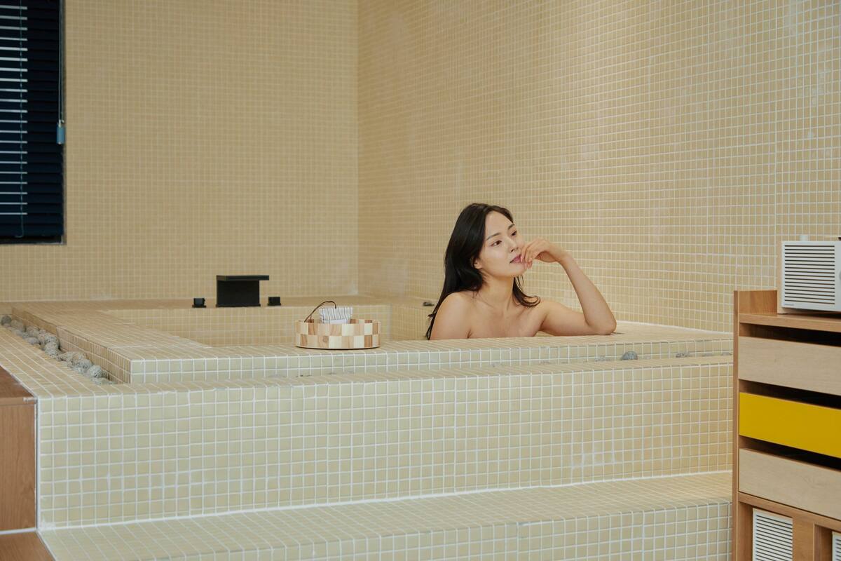 A woman relaxing in a tiled bathtub with a wooden shelf and a small wooden box on the side.