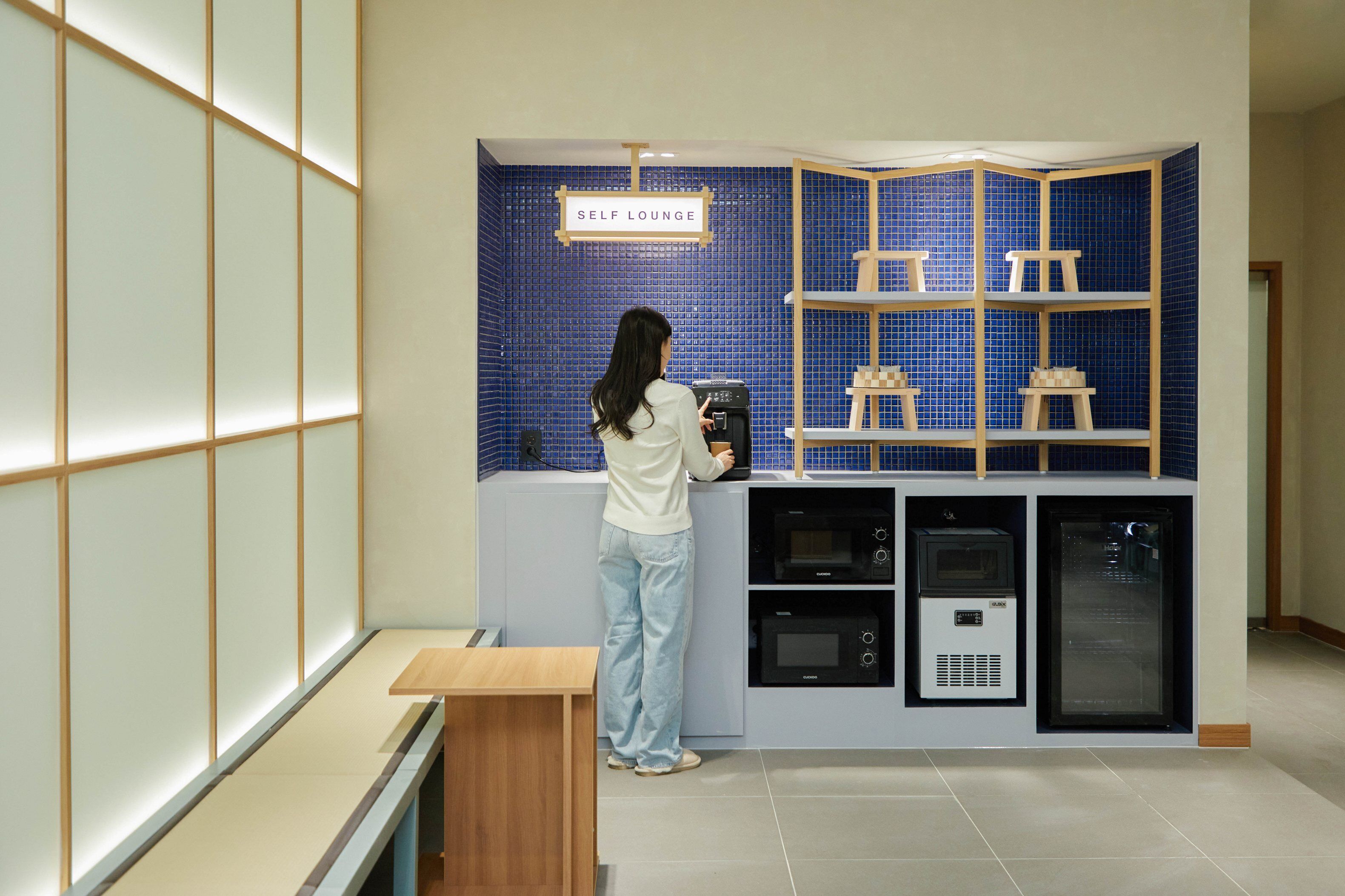 A self-lounge area with a blue tiled wall, a wooden shelf with stools, and a coffee machine.