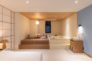 Une chambre d’hôtel moderne avec un plafond en bois, une grande baignoire, un petit coin salon et une table de chevet avec une lampe.
