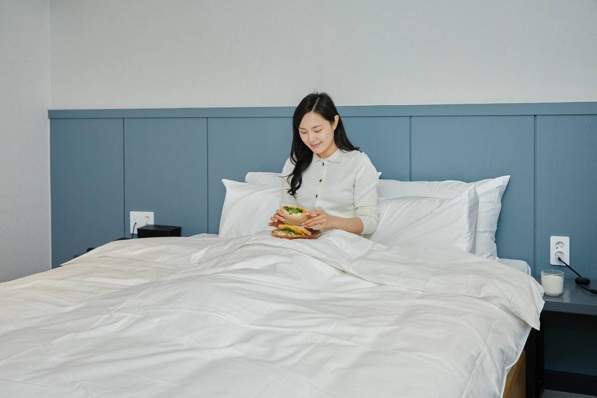 A bed with white bedding and a blue headboard, a bedside table with a lamp, and a power outlet on the wall.