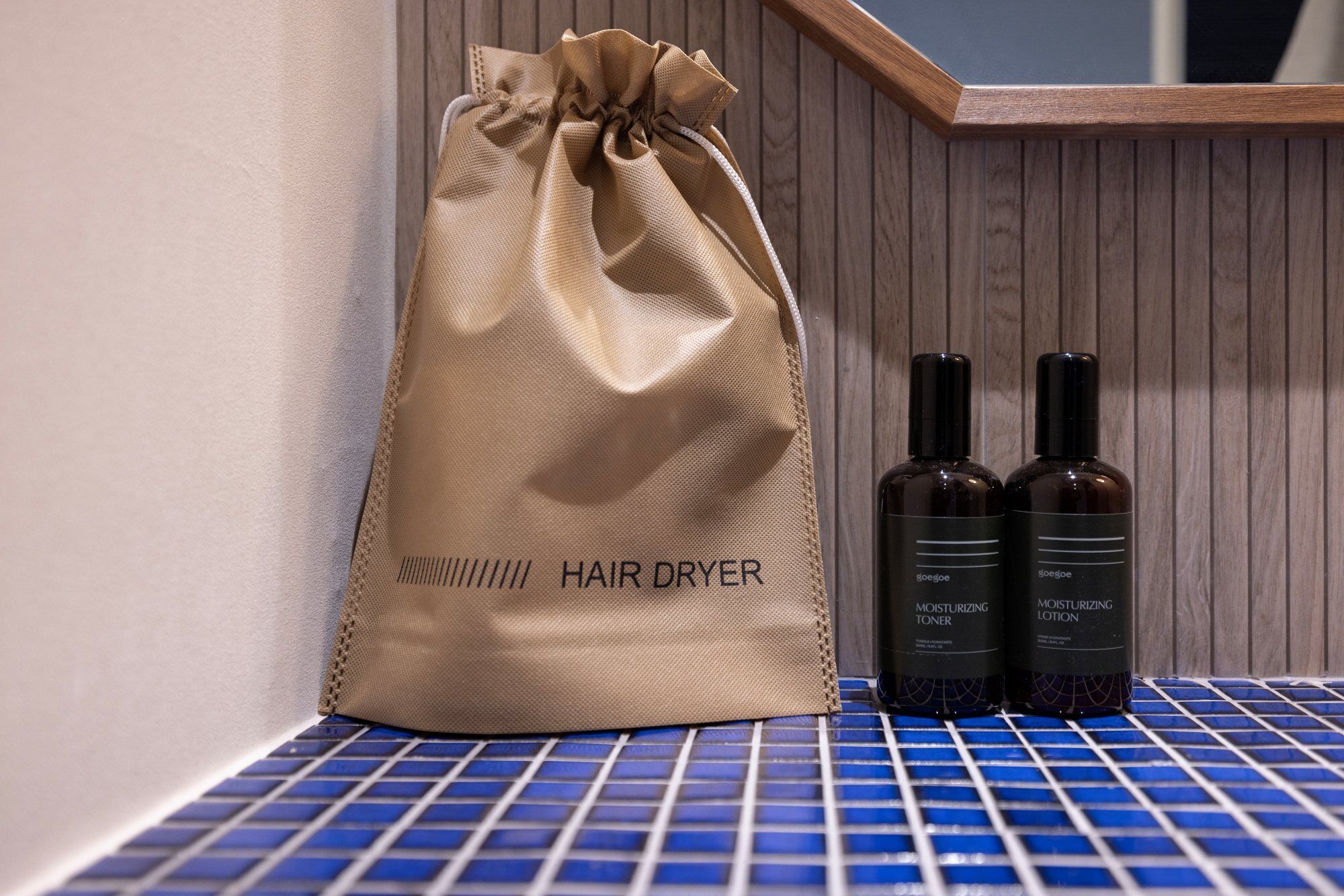 A beige drawstring bag labeled "HAIR DRYER" and two bottles of "MOISTURIZING TONER" and "MOISTURIZING LOTION" on a blue tiled surface.