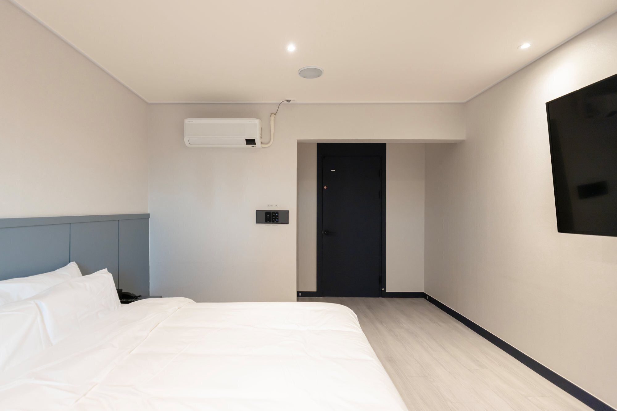 A modern hotel room with a bed, a wall-mounted TV, a control panel, and a door leading to another room.