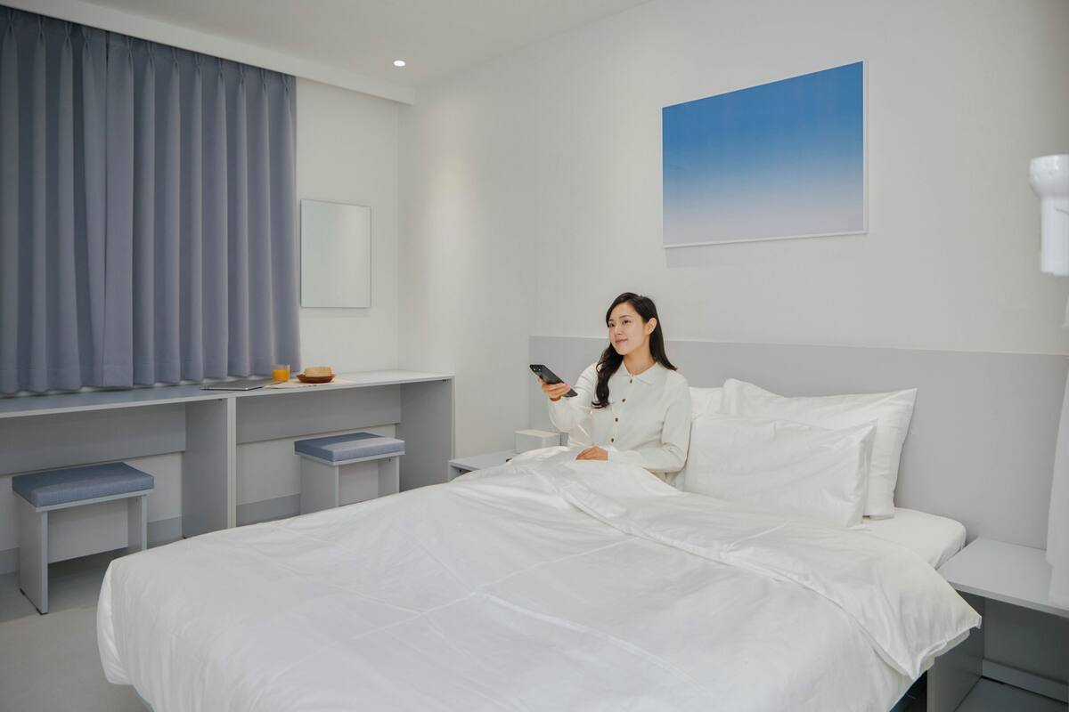 A modern hotel room with a large bed, a bedside table, a desk with a chair, and a window with curtains.