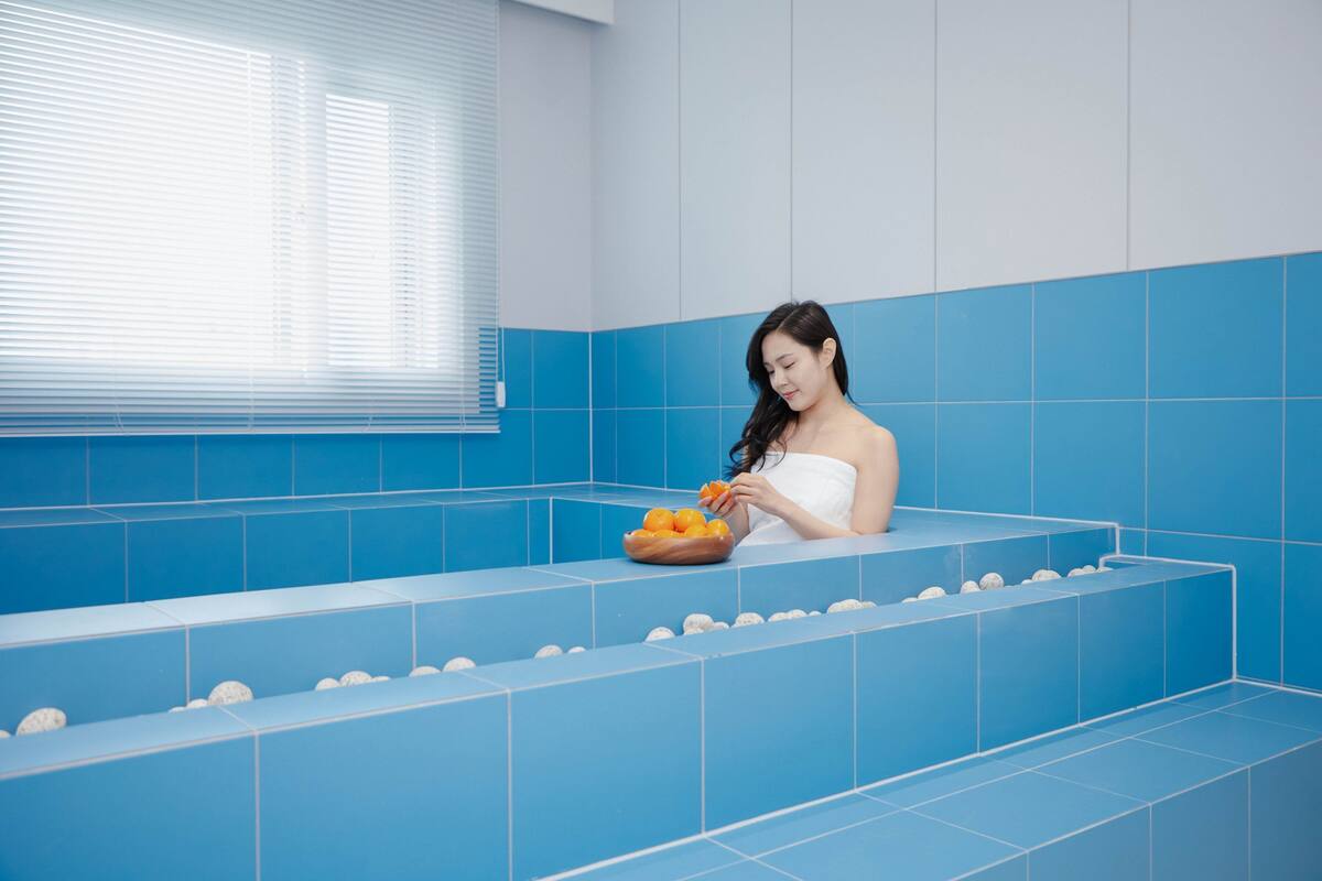 A woman in a bathtub with blue tiles, a bowl of oranges, and a window with blinds.