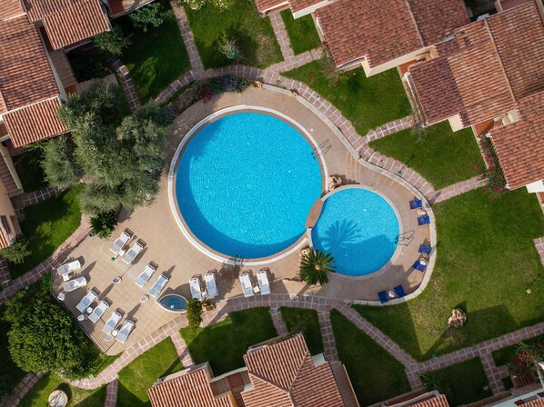 An aerial view of a resort with two large swimming pools, surrounded by lounge chairs and a well-maintained garden.