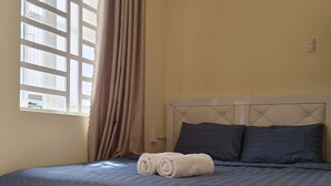 A neatly made bed with a blue bedspread and two pillows, a window with curtains, and a wall clock. - D Home (Ho Chi Minh City)