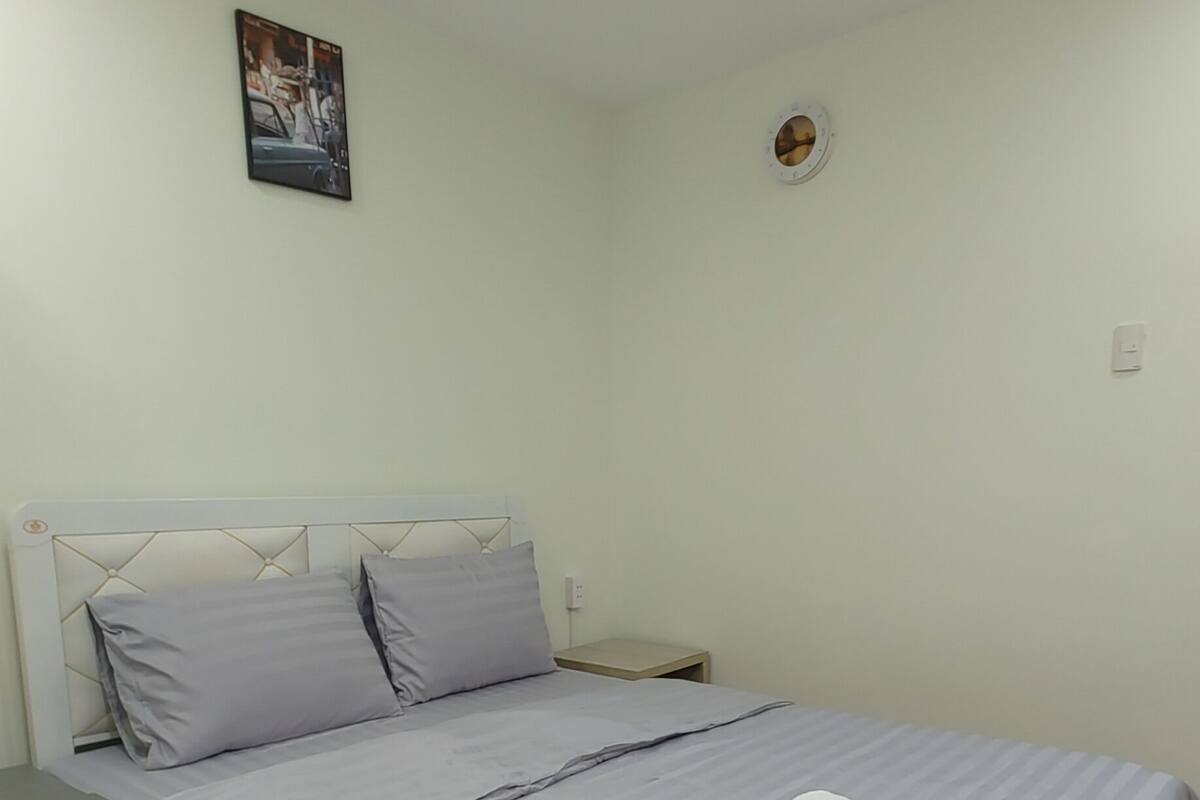 A neatly made bed with a grey bedspread and pillows, a small bedside table with a lamp, and a wall-mounted light switch.