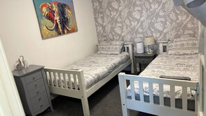 Quarto com duas camas de solteiro, mesa de cabeceira com abajur, cômoda e papel de parede estampado com flores.