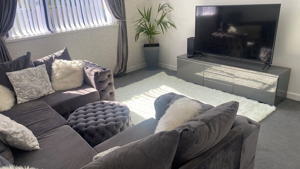 Uma sala de estar moderna com um sofá cinzento, almofadas brancas e fofas, uma televisão de tela plana grande sobre um móvel cinzento, e um vaso com planta.
