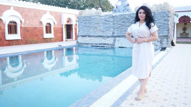 A woman standing by a pool with a stone wall and traditional architecture in the background.