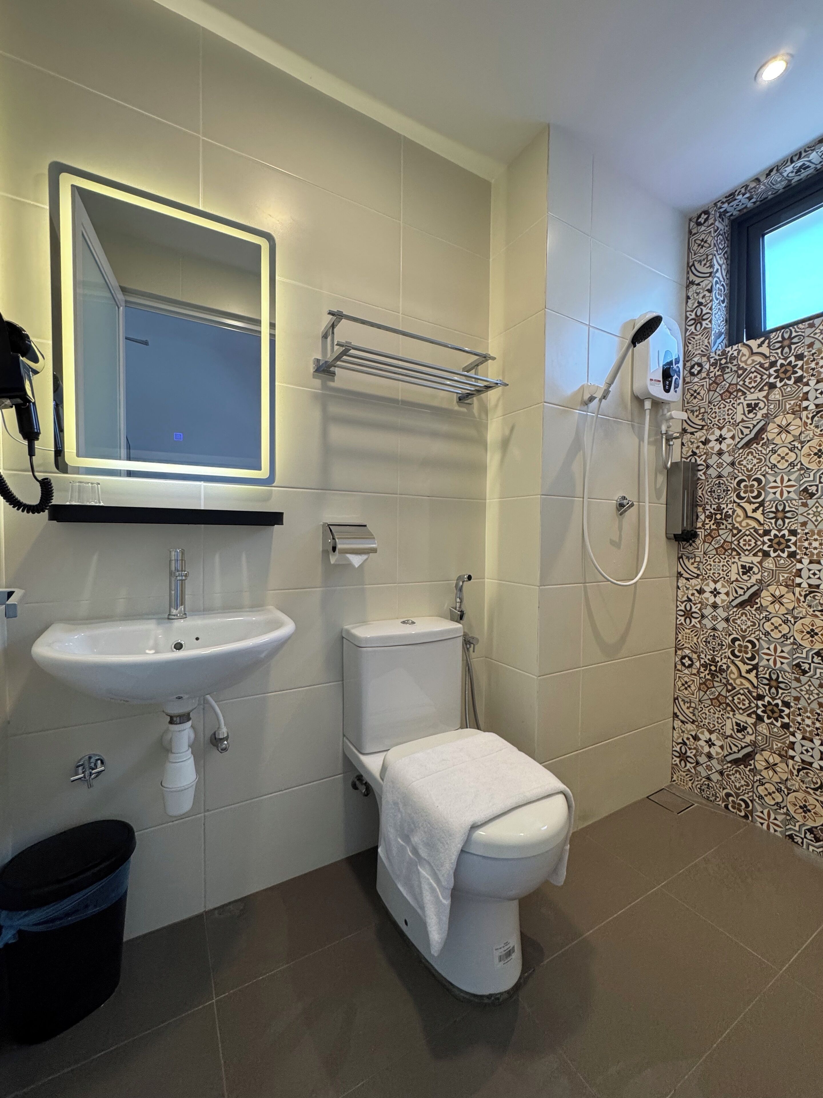 A bathroom with a white sink, toilet, and shower. There is a mirror above the sink and a towel rack above the toilet. The walls are tiled, and there is a patterned tile design on one wall.