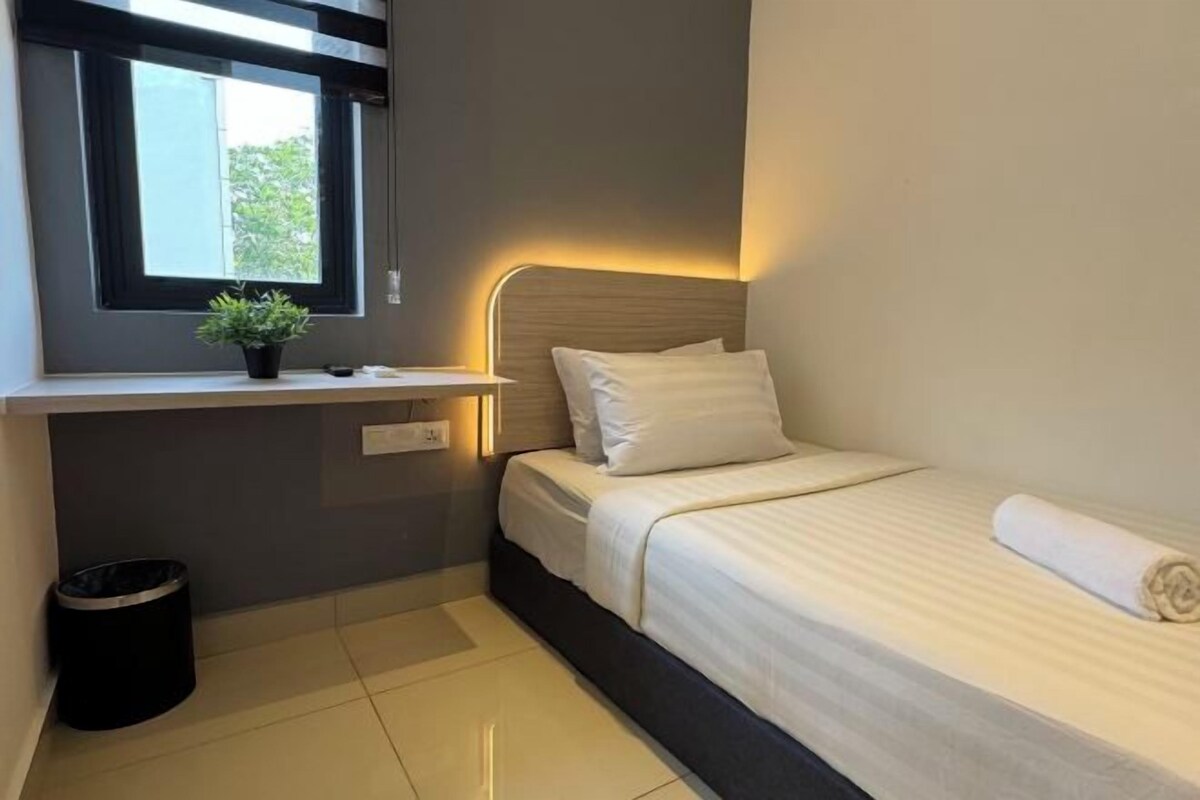 A small, neatly arranged bedroom with a single bed, a bedside table, a potted plant, and a window.