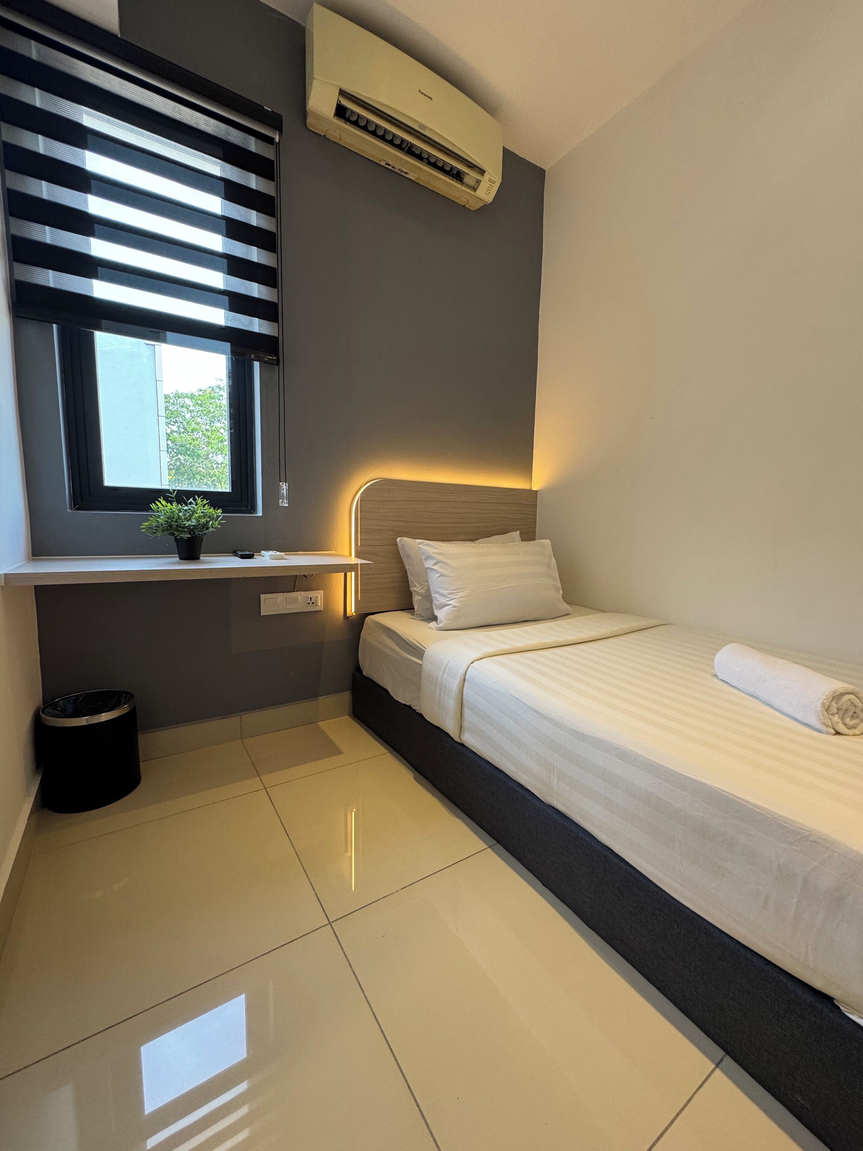 Une chambre moderne avec un lit, une fenêtre avec des stores, un climatiseur, une table de chevet avec une lampe et une petite plante en pot.