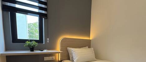 A modern bedroom with a bed, a window with blinds, an air conditioner, a bedside table with a lamp, and a small potted plant.