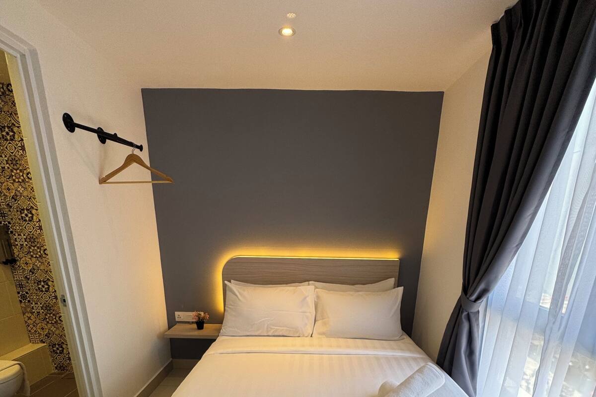 A neatly made bed with white linens, a bedside table with a lamp, and a view of the outside through sheer curtains.