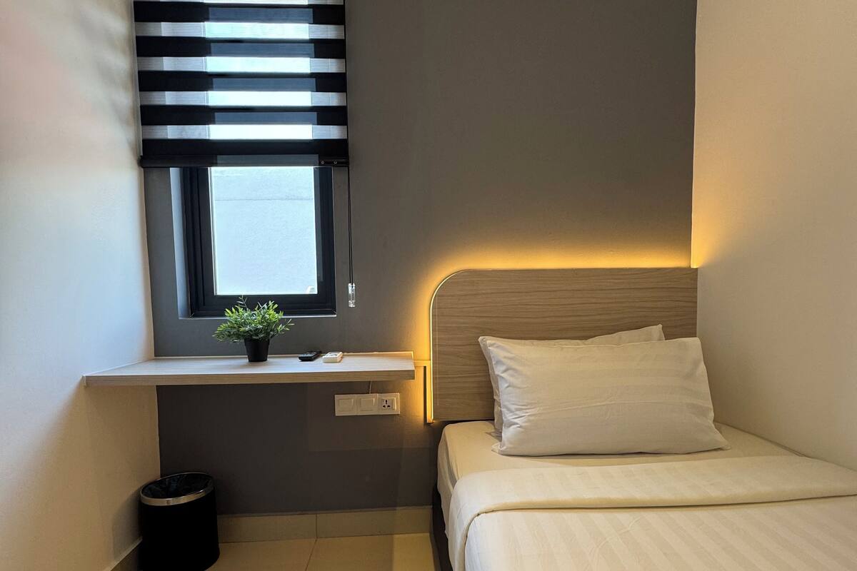 A bedroom with a bed, a window with blinds, an air conditioner, a small plant, and a trash bin.