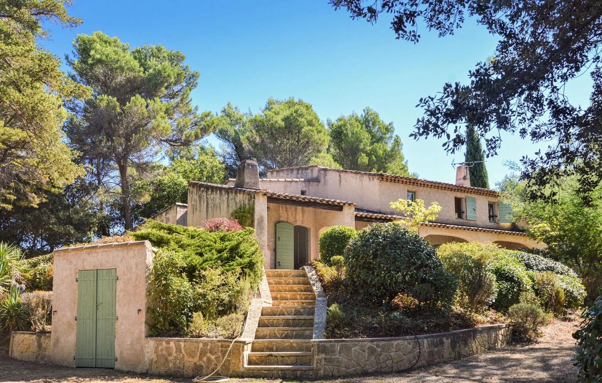 A rustic villa with a stone staircase leading to a green door, surrounded by lush greenery and trees.