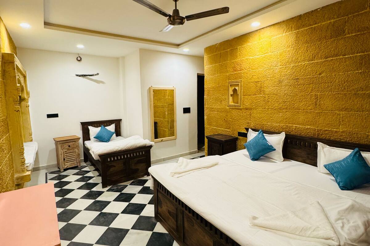 Een hotelkamer met een geruit vloertje, twee bedden met witte en blauwe kussens, een plafondventilator en een gouden wand met structuur.
