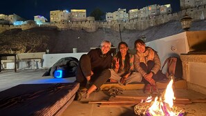 Drie personen zitten op een terras op het dak, met een vuur op de voorgrond. Achtergrond toont een historisch stenen muur met 's nachts verlichte gebouwen.