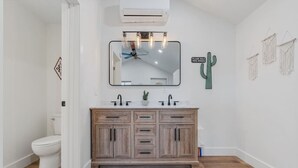 Une salle de bain avec des toilettes blanches, un meuble-lavabo en bois doté de tiroirs, un miroir avec éclairage intégré et une décoration en forme de cactus.