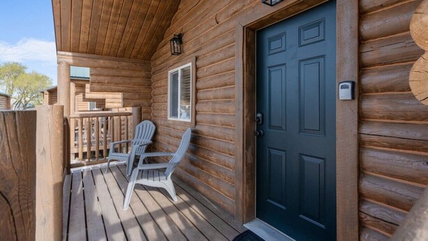Une cabane en bois avec une porte sombre, un petit porche doté de deux fauteuils Adirondack et une fenêtre avec des volets.