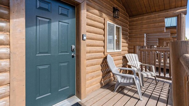 Una cabaña de troncos con una puerta azul oscuro, una ventana con contraventanas y dos sillas Adirondack en una terraza de madera.