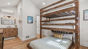 Cama tipo litera con estructura de madera, baño con lavamanos y espejo, y zona de cocina con armarios de madera.