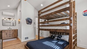 Ein Etagenbett mit Holzrahmen, eine dunkelblaue Bettdecke und ein kleines Regal darüber. An der weißen Wand sind eine Wandklimaanlage, ein Spiegel und ein gerahmtes Bild angebracht. Im Eck befindet sich ein Holzkasten mit Schubladen.