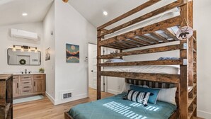 Una cama litera con estructura de madera y colcha color turquesa, un escritorio de madera con espejo, y un aire acondicionado de pared.