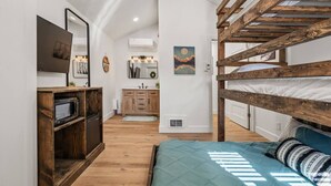 Un dormitorio con literas, piso de madera, una pequeña kitchenette y un baño con espejo y lavamanos.