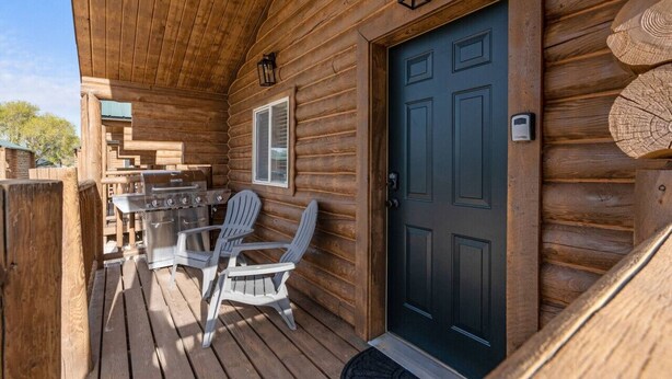 Une cabane en bois avec une véranda, un barbecue, deux fauteuils Adirondack et une porte bleu foncé.