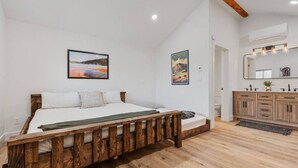 A bedroom with a wooden bed, white walls, and a wooden floor. There is a bathroom with a wooden vanity and a mirror. - #9 Yellowstone Cabin w/ AC, Fire Pit & BBQ! (Saint Charles)