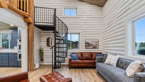 Interior de una cabaña de troncos con sala de estar, una escalera y vistas al exterior.