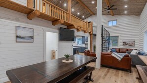 Interior espacioso de una cabaña de troncos con una mesa de comedor de madera, un sofá y una escalera que sube a una zona de altillo.