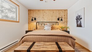 Un dormitorio con pared de listones de madera, cama con colcha acolchada, dos lámparas de noche, una mesita de noche, un banco de madera y un cuadro enmarcado en la pared.