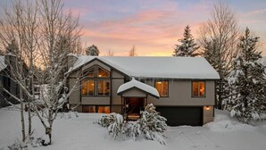 Una casa de dos pisos con tejado nevado y entrada, rodeada de árboles y un cielo al atardecer.