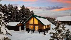 A log cabin with a snow-covered roof, surrounded by snow-laden trees, under a twilight sky.