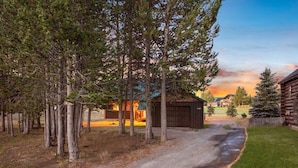 A gravel driveway leading to a wooden cabin with a garage, surrounded by trees and a grassy area.