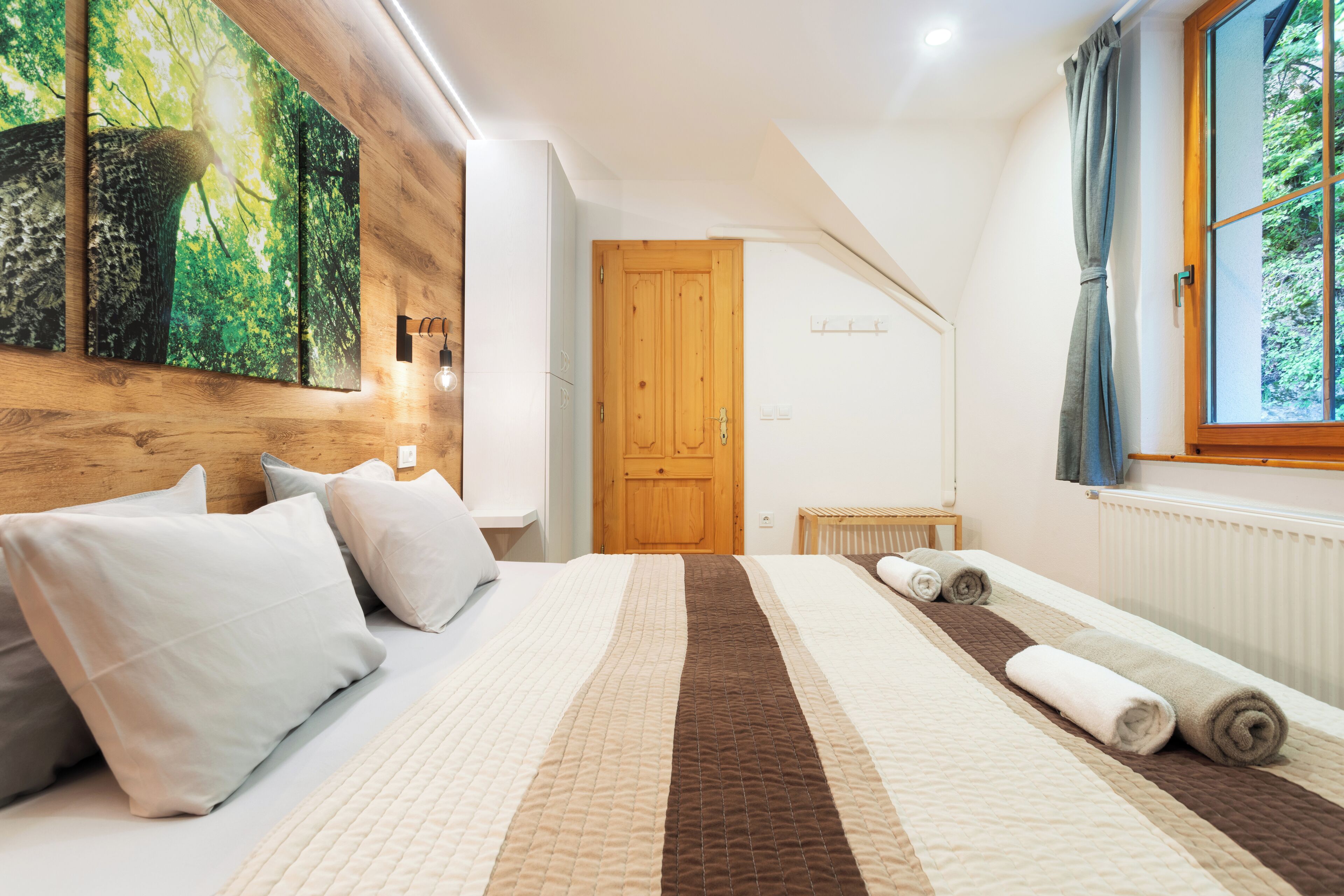 A bedroom with a large bed, a wooden door, a window with curtains, and a wall-mounted light fixture.