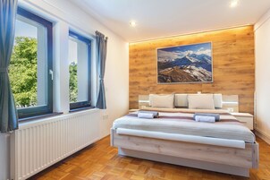 Un dormitorio con cabecera de madera, un ventanal con cortinas, una cama con ropa de cama blanca y un cuadro de un paisaje montañoso en la pared.