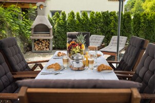 Un espacio de comedor al aire libre con una parrilla de piedra, un tarro de frutas y galletas, cruasanes y una mesa lista para dos.