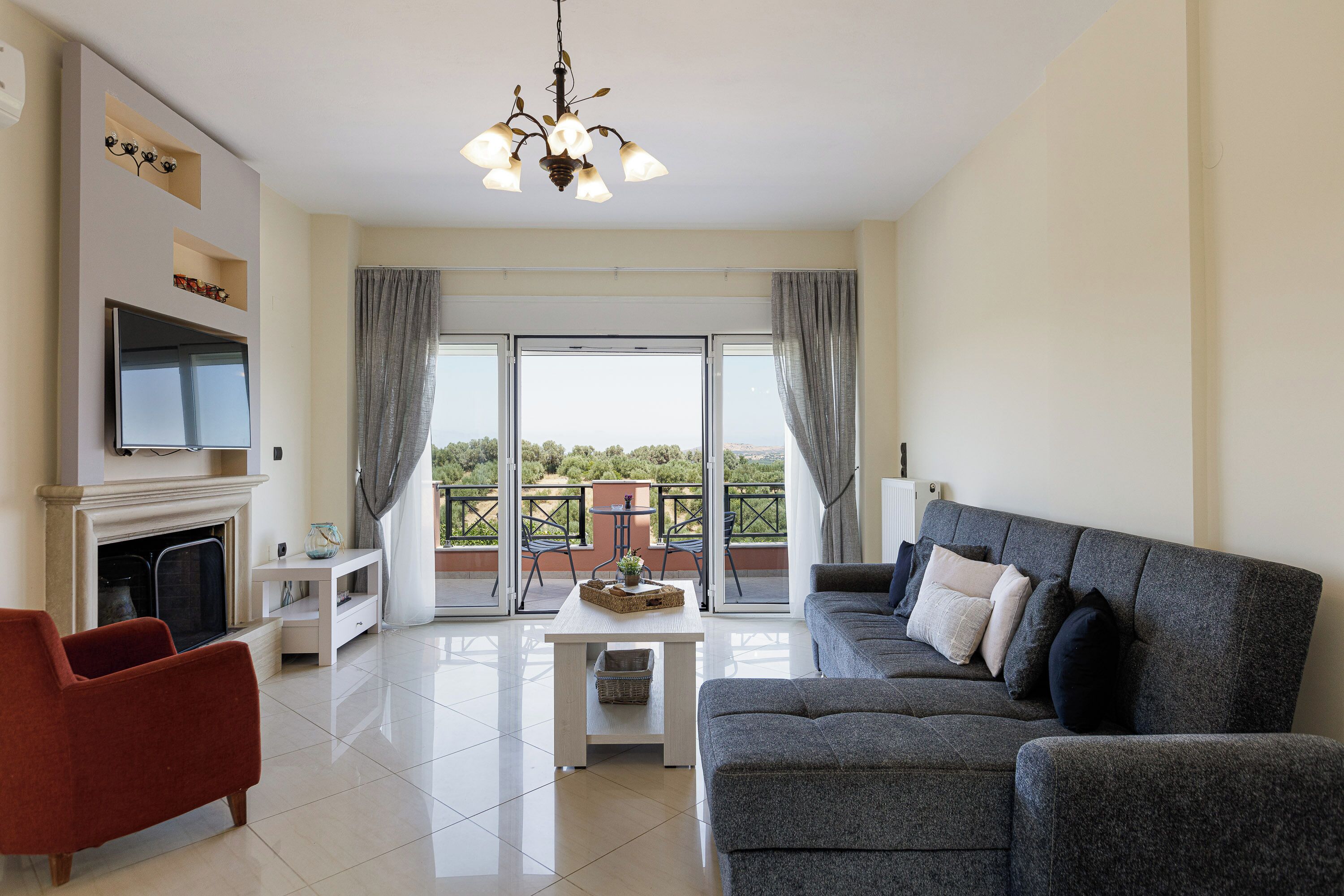 A modern living room with a fireplace, a grey sofa, a red armchair, and a balcony with a view.