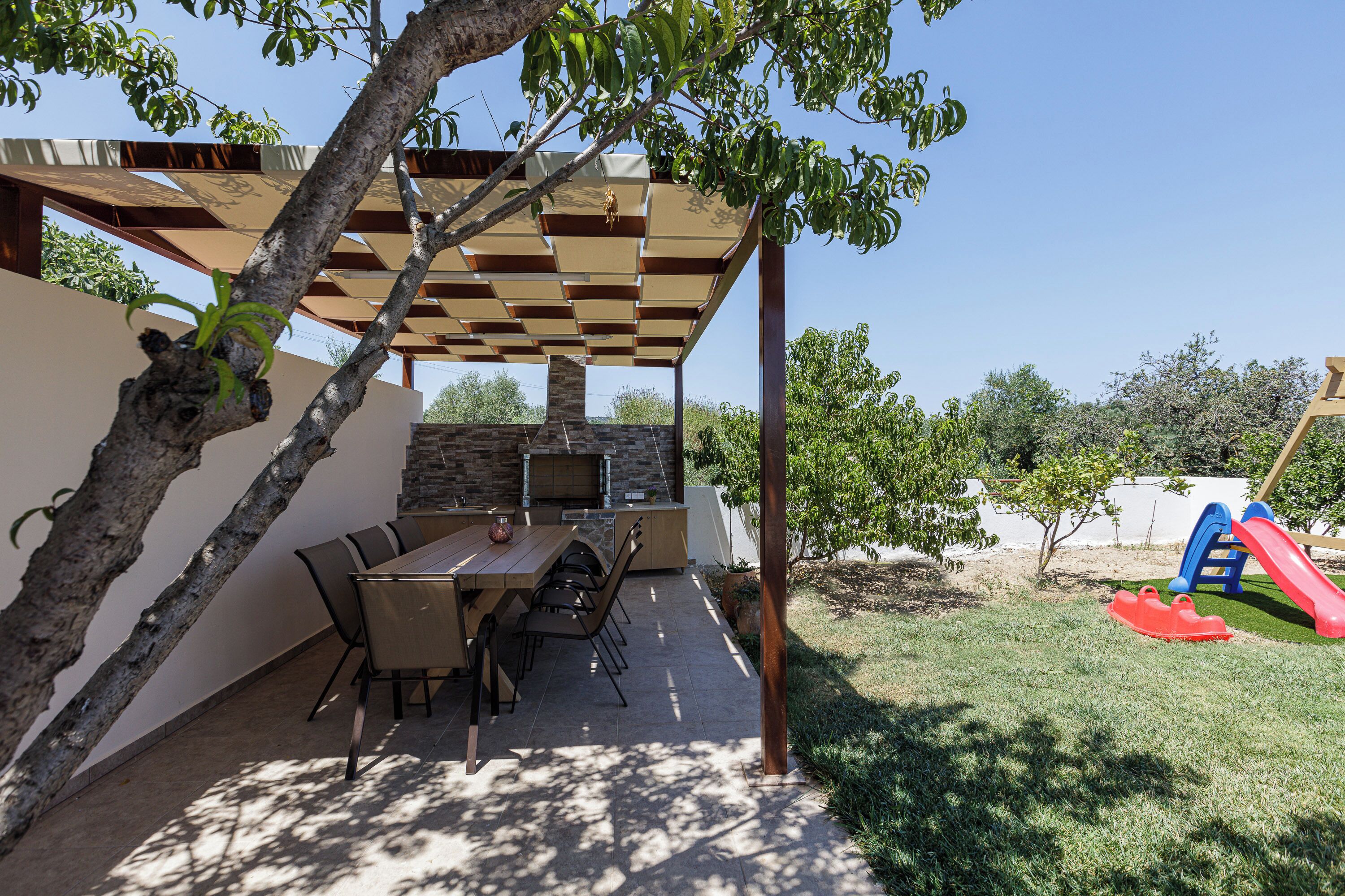 Un patio exterior con mesa y sillas, una chimenea de piedra y una zona de juegos infantiles con un tobogán.