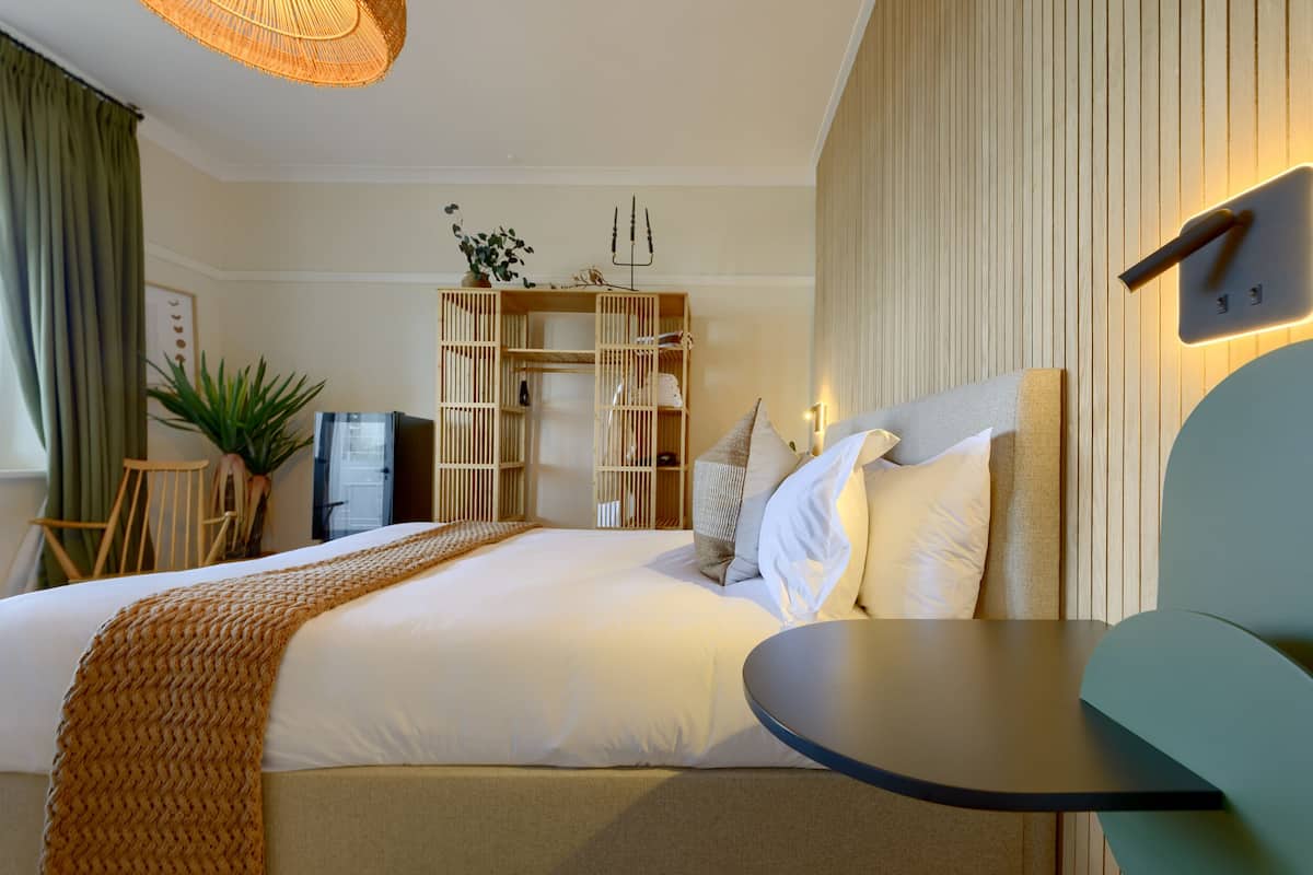 A hotel room with a large bed, a small table, a bookshelf, and a wall-mounted light fixture.