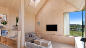 A modern living room with a sloped ceiling, built-in wooden shelving, a flat-screen TV, and a large window offering a view of the outdoors. - 5 Star Holiday Home in Lokken (Lokken)