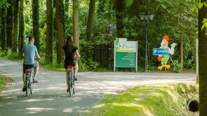 Dos personas andan en bicicleta por un camino pavimentado rodeado de árboles. Hay un cartel que indica 'Ruwinkel' y una señal de estacionamiento con una 'P' y un pato como mascota.