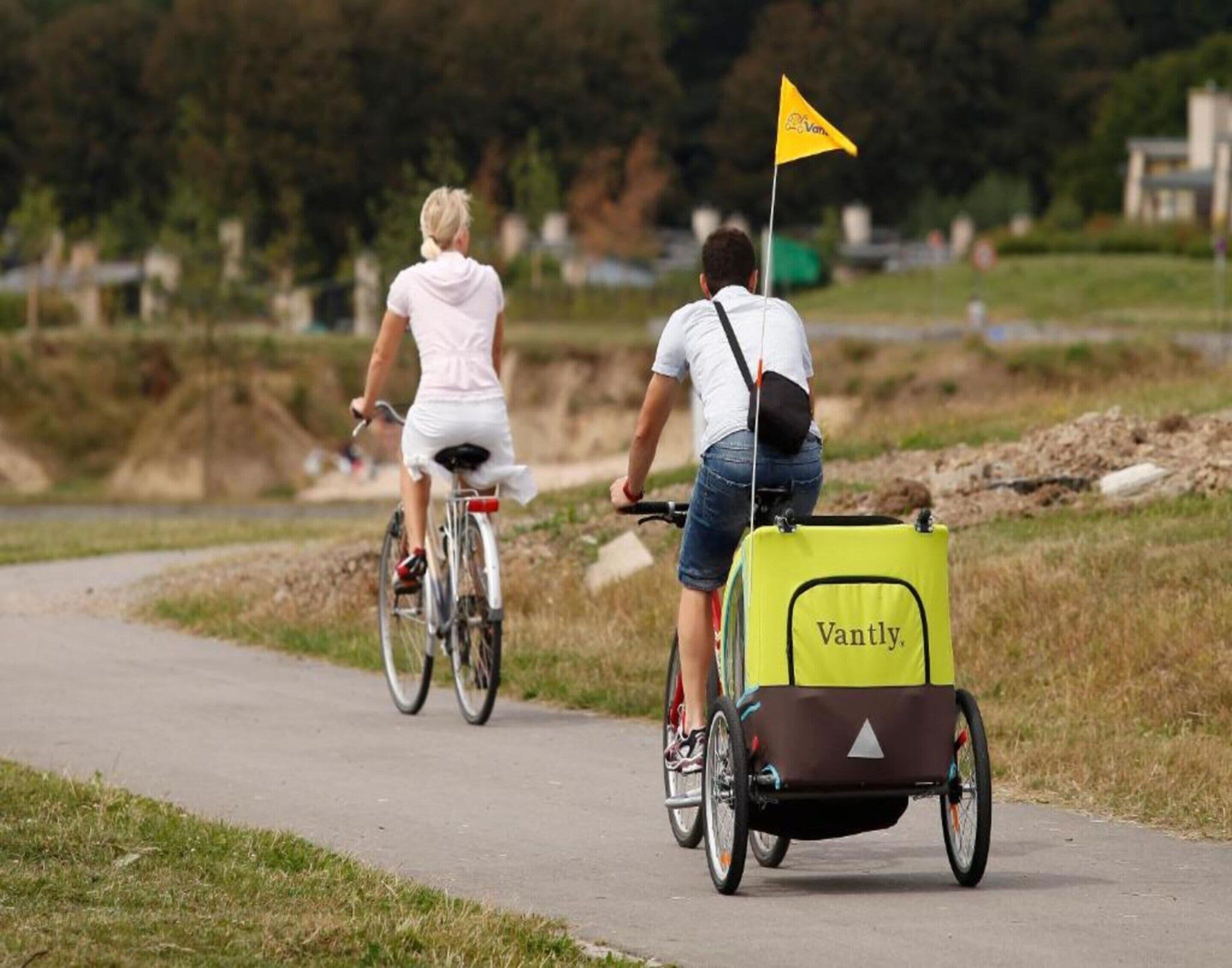 Twee personen fietsen op een verhard pad met een gele vlag, waarvan er één een gele bezorgdoos draagt met de tekst 'Vantly'.