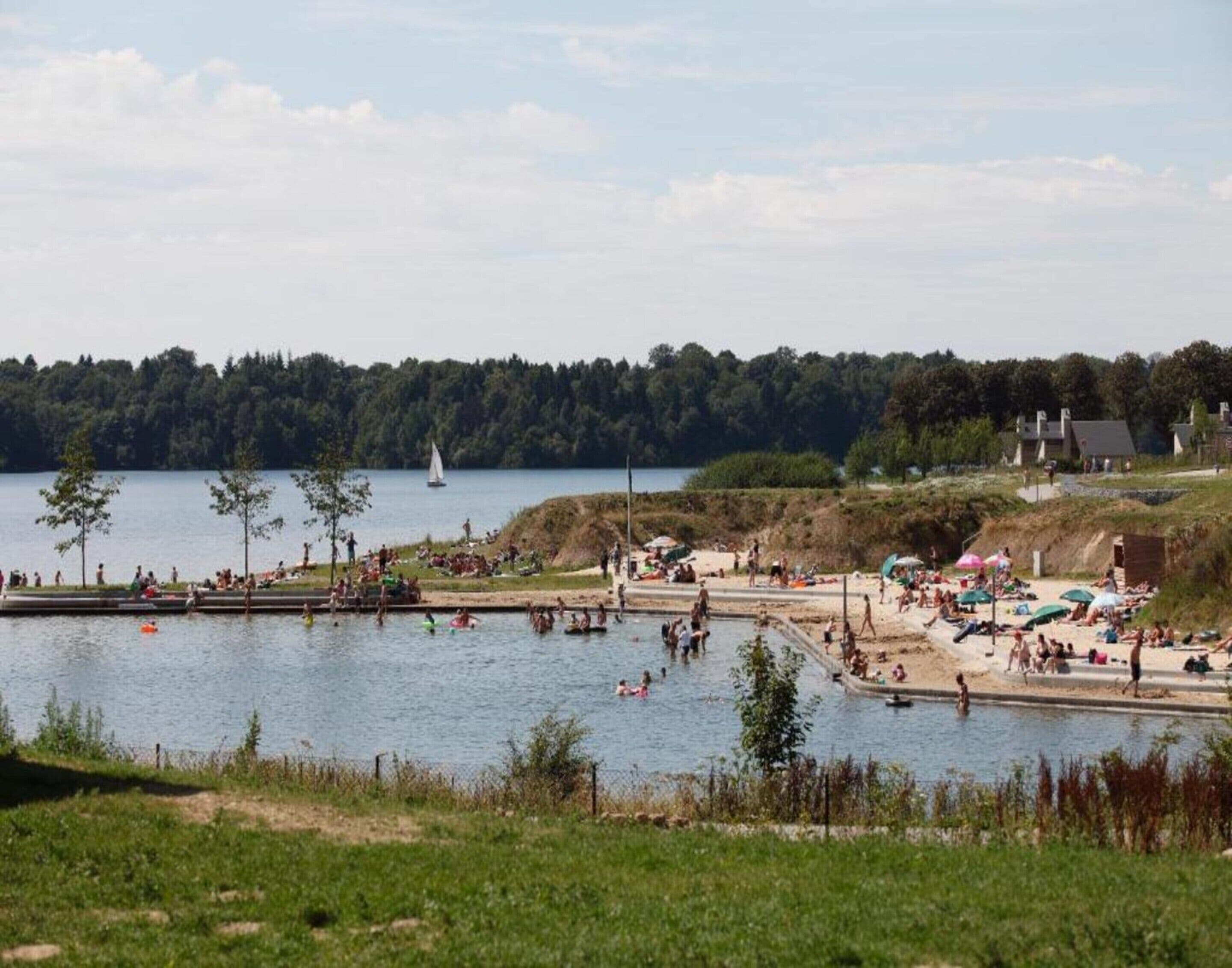 A lakeside beach with people swimming and sunbathing, a sailboat on the water, and a building in the background.